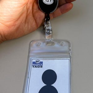 May include: A clear plastic badge holder with a black and white employee badge inside. The badge has a silhouette of a person and the text "EMPLOYEE". The badge holder is attached to a retractable badge reel with a black oval and the number "13.1" printed on it.