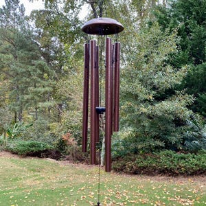 May include: Bronze-colored wind chimes hanging outdoors. The chimes have a circular top and bottom, with several long, cylindrical tubes suspended from the top. The background features green trees and grass.