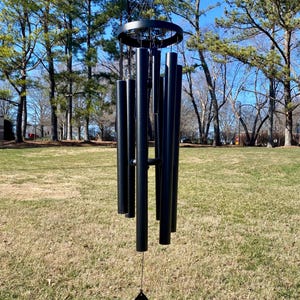 Puede incluir: Carillón de metal negro con un badajo en forma de lágrima que cuelga de un marco circular. El carillón tiene varios tubos de diferentes longitudes.