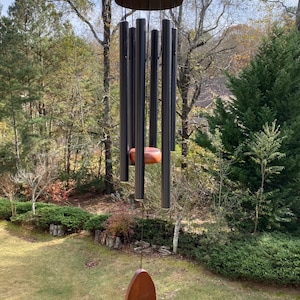 May include: A wind chime featuring a circular wooden top and a matching wooden striker suspended by black cords. Six matte black metal tubes of varying lengths hang from the top, with a teardrop-shaped wooden wind catcher at the bottom.