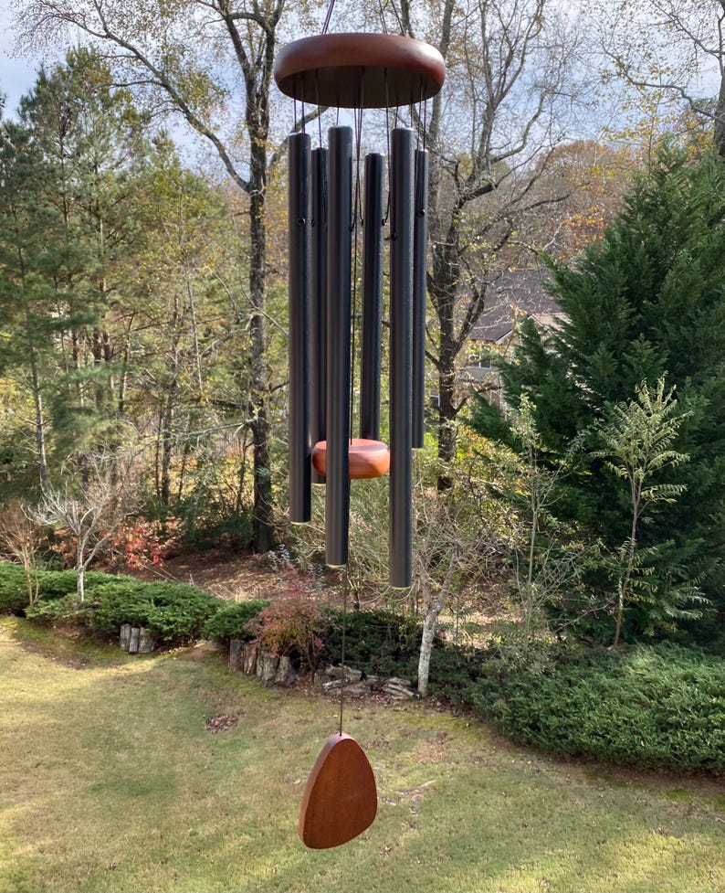 May include: A wind chime featuring a circular wooden top and a matching wooden striker suspended by black cords. Six matte black metal tubes of varying lengths hang from the top, with a teardrop-shaped wooden wind catcher at the bottom.