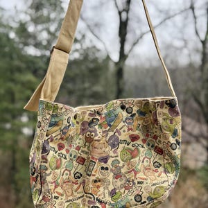 May include: A beige canvas messenger bag with a colorful cartoon character print. The bag has a long, adjustable strap and a flap closure. The print features various monsters, aliens, and other quirky characters with text like "Stay Cool" and "Wow!"