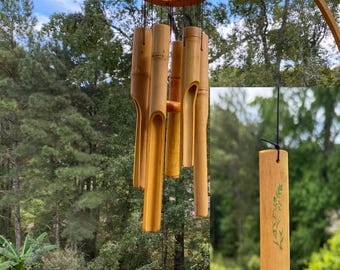 May include: Bamboo wind chimes with a natural finish. The chimes have a wooden ring at the top and several bamboo tubes hanging below. The tubes are varying lengths and have cut-out sections. The background shows a lush green outdoor setting.