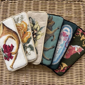 Può includere: Una collezione di sei astucci rettangolari in mezzo punto con vari disegni, tra cui un corno dorato con fiori rossi, fiori gialli, un girasole, un cervo che salta, un motivo astratto blu e una volpe su uno sfondo verde scuro.