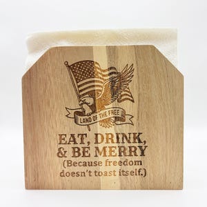 Pode incluir: Porta-guardanapos de madeira com uma bandeira americana e uma águia gravadas. O texto diz "LAND OF THE FREE" e "EAT, DRINK, & BE MERRY (Because freedom doesn't toast itself.)". Contém guardanapos brancos.