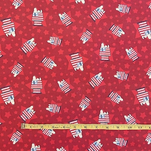 May include: Red fabric with a repeating pattern of Snoopy in a house decorated with the American flag, set against a background of red stars. The fabric is measured with a yellow ruler.