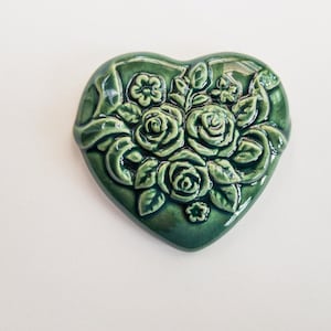 May include: A heart-shaped ceramic object with a glossy dark green glaze. The surface features a raised, molded relief design of three central roses surrounded by smaller flowers, leaves, and swirling ribbon-like accents in the same green tone.