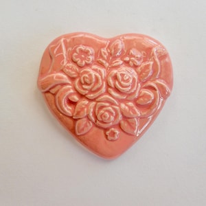 May include: A heart-shaped, ceramic ornament in a soft coral color. The ornament features a raised floral design with roses and leaves. The details are highlighted with a lighter shade of coral, creating a three-dimensional effect.