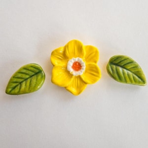 May include: A ceramic daffodil with a bright yellow flower, an orange center, and two green leaves. The flower and leaves are arranged on a white surface. The daffodil is a decorative item.