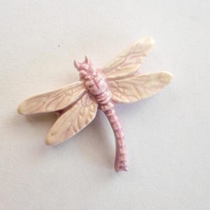 May include: A detailed, light pink dragonfly brooch with cream-colored wings. The dragonfly has a textured body and wings, and is set against a white background. This decorative pin is a nature-inspired accessory.