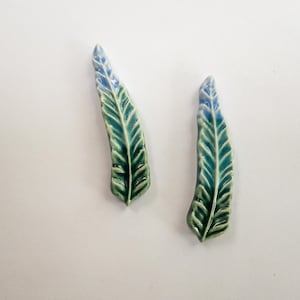 May include: Two ceramic leaf-shaped decorative pieces. The leaves are a mix of teal and green with blue tips and detailed veining. They are placed on a white background.