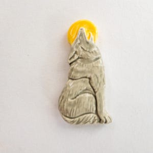 May include: A ceramic figurine of a howling wolf with a yellow moon behind its head. The wolf is light gray with carved details, and the moon is a bright yellow. The figurine is likely a decorative item or a collectible.
