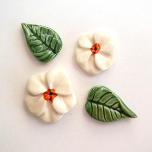 May include: Two ceramic flowers and two ceramic leaves. The flowers are off-white with five rounded petals and a bright orange center with a black dot. The leaves are green with a glossy finish and visible dark green veining throughout the surface.