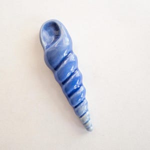 May include: A blue ceramic seashell sculpture with a spiral design. The sculpture is painted in shades of blue, with darker blue stripes along the spiral. The seashell has a smooth, glossy finish and is a decorative item.