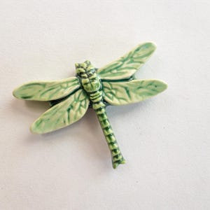 May include: A ceramic dragonfly figurine in shades of green. The dragonfly has detailed wings and body, with a textured appearance. The wings are a lighter green, while the body is a darker green. The figurine is a decorative item.