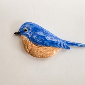 May include: A ceramic bluebird figurine with a glossy finish. The bird has a blue body, a tan breast, and a black beak. The figurine is designed to resemble a real bird.