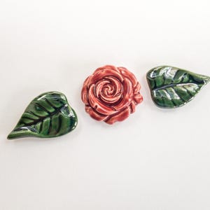 May include: Ceramic rose and leaf set. The rose is a deep red with detailed petals. Two green leaves with dark veins flank the rose. The pieces are likely decorative or for crafts.