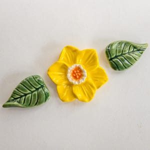 May include: A ceramic daffodil with a bright yellow flower and a white and orange center, accompanied by two green ceramic leaves. The handmade floral decor is set against a white background.