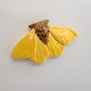 May include: A ceramic moth figurine with bright yellow wings and a brown body. The wings are detailed with subtle ridges, and the body has a textured appearance. The figurine is set against a plain white background.