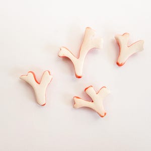 Puede incluir: Cuatro peque&ntilde;os adornos con forma de coral ramificado en color crema p&aacute;lido con bordes de color naranja rojizo vibrante. Cada pieza presenta una estructura en forma de Y con m&uacute;ltiples puntas, un acabado mate liso y formas org&aacute;nicas.