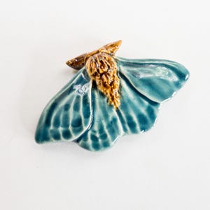May include: A ceramic moth figurine with teal wings and a brown body. The wings have detailed veining and a glossy finish. The moth is a decorative item, likely for display or collection.