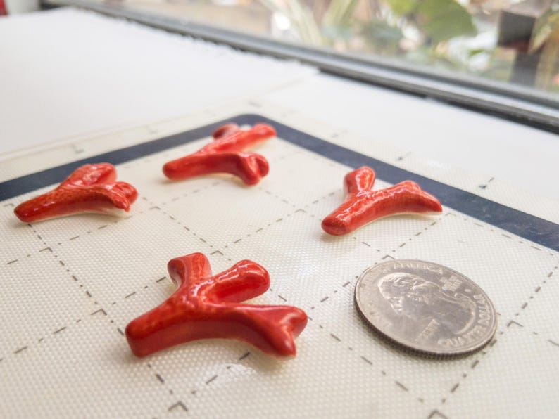 Puede incluir: Cuatro piezas peque&ntilde;as de cer&aacute;mica de color rojo brillante con forma de fragmentos de coral ramificados. Las piezas est&aacute;n dispuestas sobre una alfombrilla de rejilla blanca junto a una moneda plateada para comparar el tama&ntilde;o y textura.