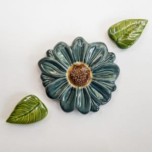 May include: A ceramic wall art set consisting of one large flower with dark teal petals and a textured brown center, accompanied by two separate green leaves with visible vein details, all featuring a glossy finish and rounded edges.