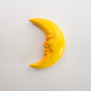 May include: A yellow crescent moon with a face carved into it. The moon is a bright, sunny yellow color and has a glossy finish. The face has detailed features, including a nose, mouth, and eyes. The moon is a decorative item.