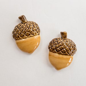May include: Two ceramic acorn ornaments. The top of each acorn is a dark brown color with a textured pattern, while the bottom is a light tan color. The acorns are set against a white background.