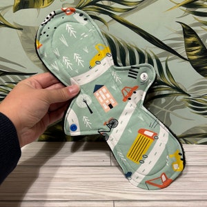 May include: A green reusable cloth menstrual pad with a black trim and a whimsical print of cars, houses, and trees. The pad has blue snap buttons on the sides.