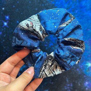 May include: A blue and white fabric scrunchie with a Star Wars spaceship print. The scrunchie is made of soft, stretchy fabric and is perfect for holding hair in place.