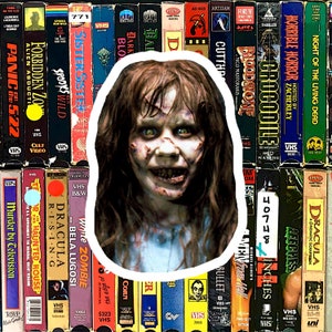Puede incluir: Primer plano de una niña joven con cabello largo y oscuro y un vestido blanco. Su rostro es pálido y sus ojos están muy abiertos con una sonrisa siniestra. La imagen está rodeada por una forma ovalada blanca y un fondo de cintas VHS.