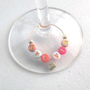 May include: A gold-colored wine glass charm with the name "SYLVIA" spelled out in colorful beads. A small gold heart charm hangs from the bottom of the charm.