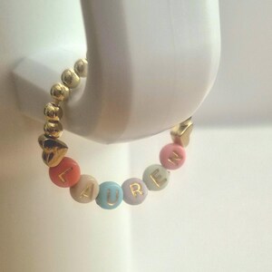 May include: A gold beaded bracelet with the name "LAUREN" spelled out in colorful letter beads. The bracelet is held by a white object. The bracelet has gold heart-shaped accents.