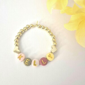 May include: Gold beaded bracelet with the name "KELLY" spelled out in colorful letter beads. The bracelet includes gold heart-shaped charms and is set against a white background with yellow flowers.