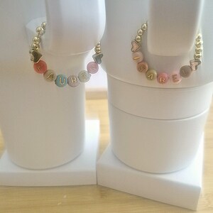 May include: Two personalized bracelets, each featuring a name spelled out with colorful letter beads. The bracelets have gold-colored beads and heart or star-shaped accents. One bracelet spells "LAUREN" and the other spells "ISABEL".