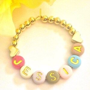 May include: A gold beaded bracelet with colorful letter beads spelling "Jessica". The bracelet features gold heart-shaped charms and is set against a white background with a yellow flower.