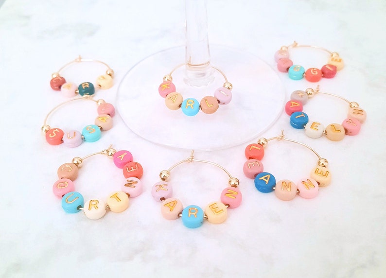 May include: Set of 8 gold-toned wine glass charms with colorful beads spelling out names. The names are: Courtney, Karen, Anne, Lexi, Layla, Karl, Susan, and Ginger.