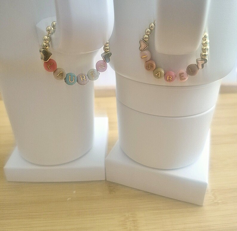 May include: Two personalized bracelets, each featuring a name spelled out with colorful letter beads. The bracelets have gold-colored beads and heart or star-shaped accents. One bracelet spells "LAUREN" and the other spells "ISABEL".