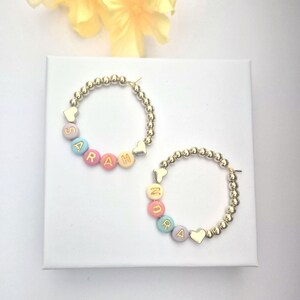 May include: Two gold-colored hoop earrings with beaded detailing. One earring spells "SARAH" with pastel-colored letter beads. The other earring spells "MORA". Each earring has a heart-shaped charm. The earrings are on a white surface.