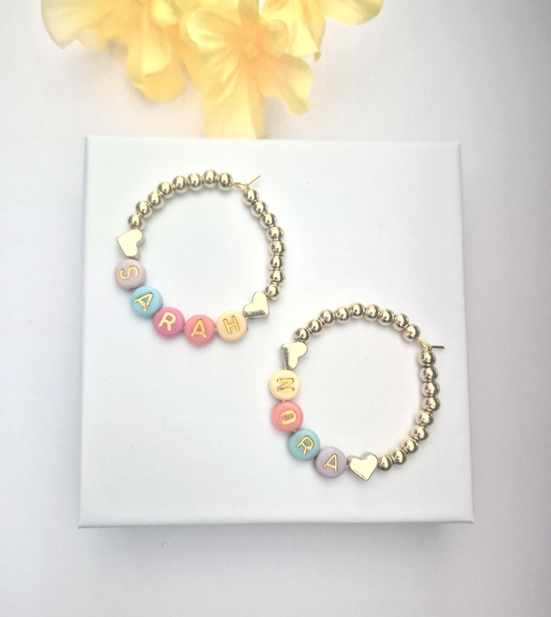May include: Two gold-colored hoop earrings with beaded detailing. One earring spells "SARAH" with pastel-colored letter beads. The other earring spells "MORA". Each earring has a heart-shaped charm. The earrings are on a white surface.