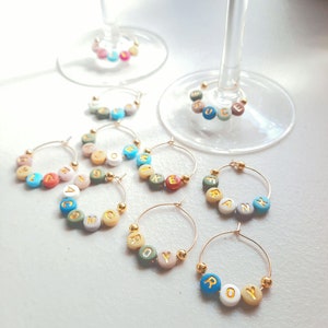 May include: Six gold-toned wine glass charms with colorful beads spelling out the names "COM", "ROY", "FRANK", "MIKE", "ANNA", and "JUC".