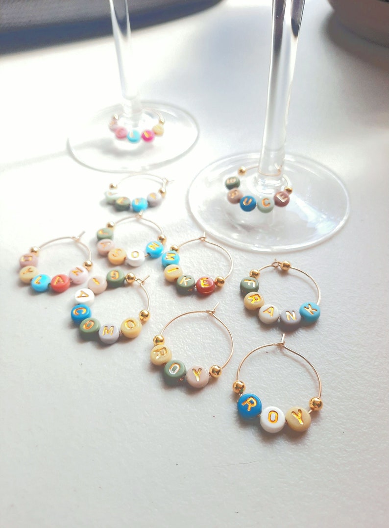 May include: Six gold-toned wine glass charms with colorful beads spelling out the names "COM", "ROY", "FRANK", "MIKE", "ANNA", and "JUC".