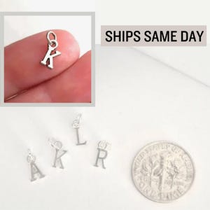 May include: Small silver-toned metal letter charms in the shapes of A, K, L, and R, each featuring a circular jump ring at the top for attachment. A United States dime coin is positioned next to the charms to provide a visual scale reference.