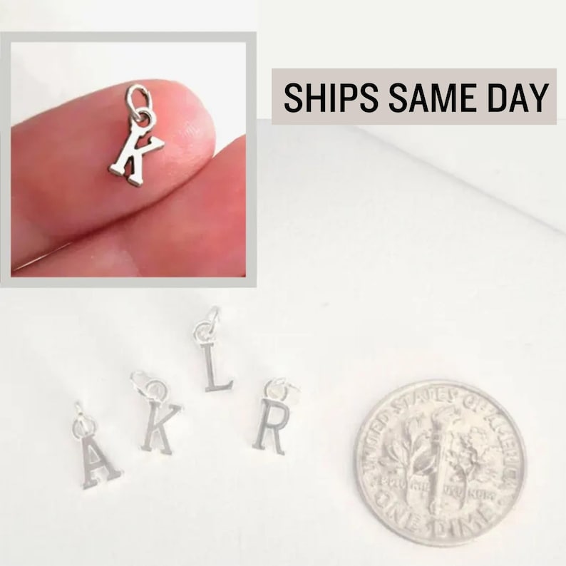 May include: Small silver-toned metal letter charms in the shapes of A, K, L, and R, each featuring a circular jump ring at the top for attachment. A United States dime coin is positioned next to the charms to provide a visual scale reference.