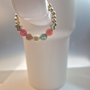 May include: A gold beaded bracelet spelling "JENNY" with colorful letter beads and heart-shaped accents. The bracelet is displayed on a white background. The bracelet is a personalized jewelry item.