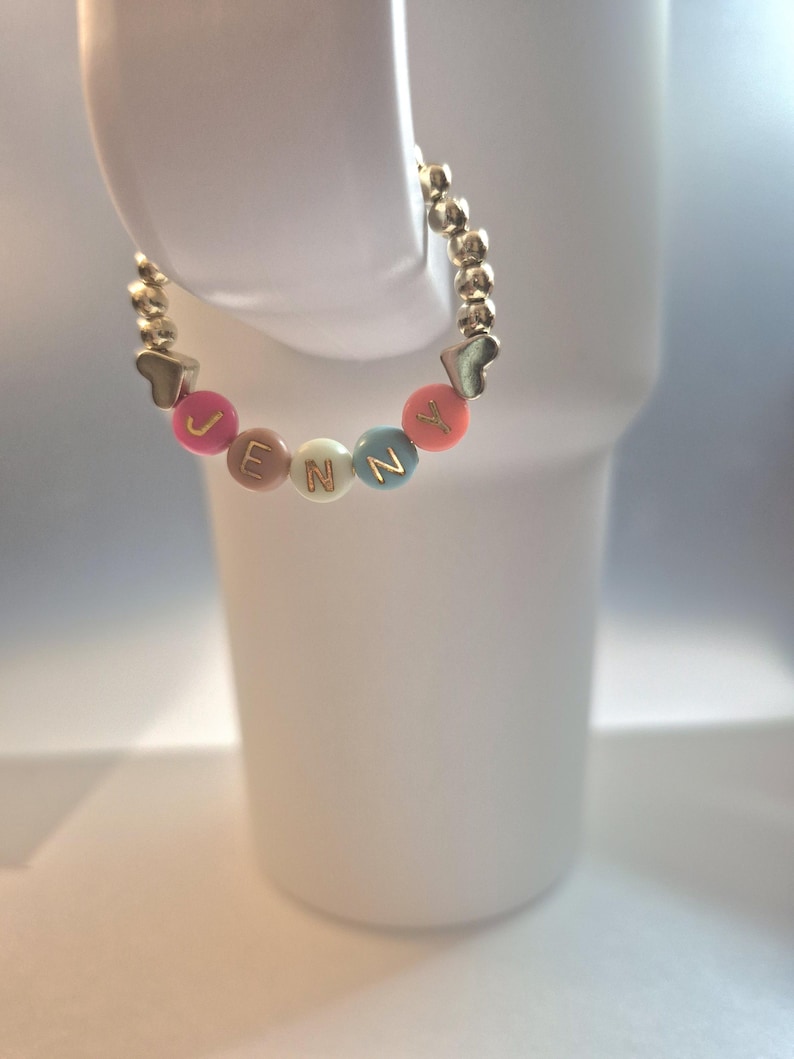 May include: A gold beaded bracelet spelling "JENNY" with colorful letter beads and heart-shaped accents. The bracelet is displayed on a white background. The bracelet is a personalized jewelry item.
