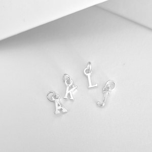 May include: Four small silver-toned metal letter charms featuring the uppercase letters A, K, L, and J. Each individual letter charm is attached to a small circular jump ring, designed for use as a component in jewelry making or decorative accessories.