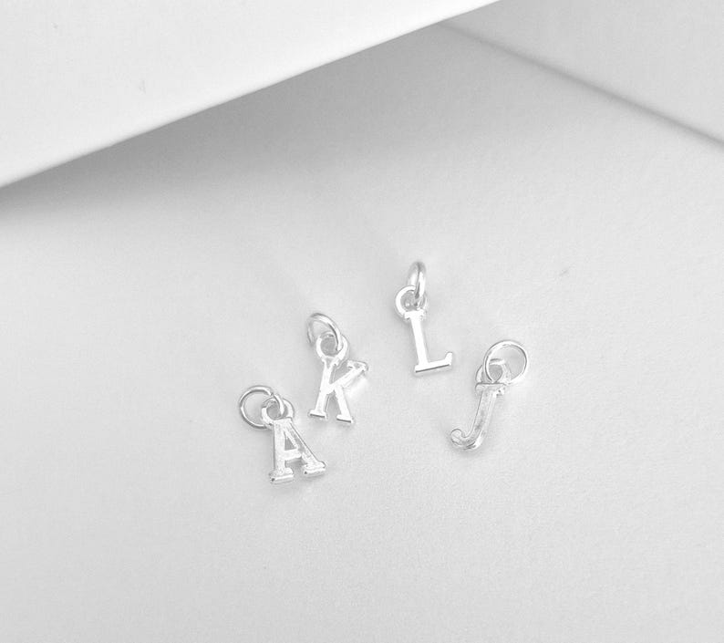 May include: Four small silver-toned metal letter charms featuring the uppercase letters A, K, L, and J. Each individual letter charm is attached to a small circular jump ring, designed for use as a component in jewelry making or decorative accessories.