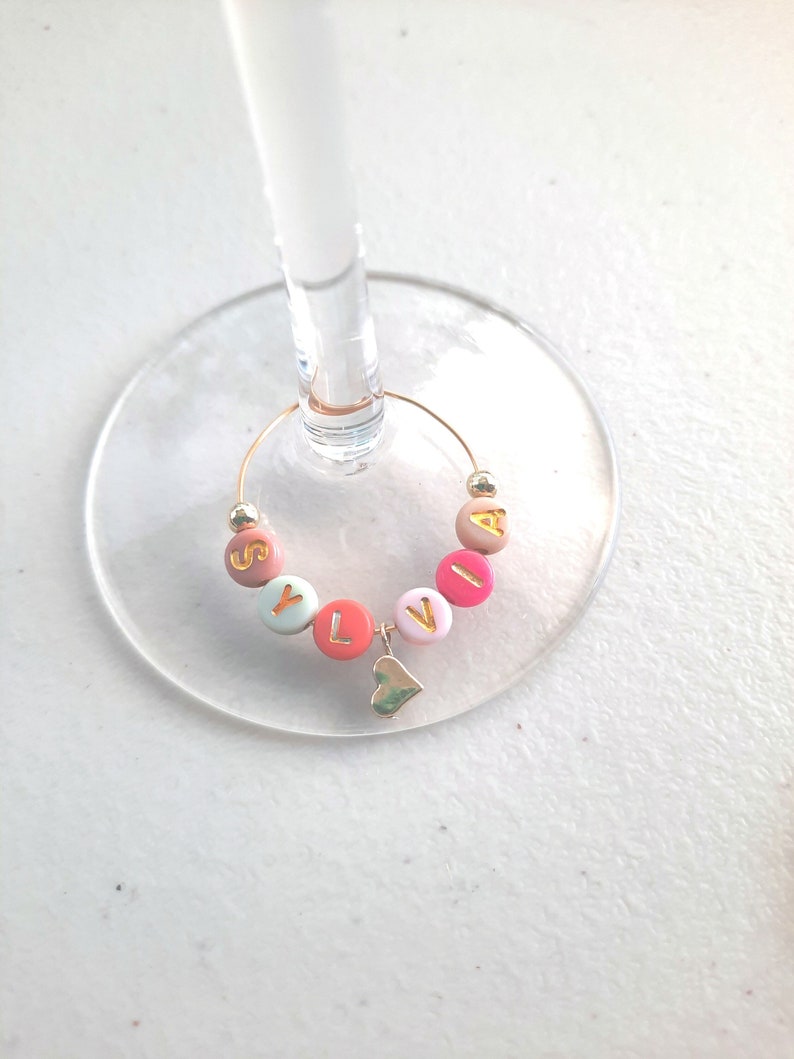 May include: A gold-colored wine glass charm with the name "SYLVIA" spelled out in colorful beads. A small gold heart charm hangs from the bottom of the charm.